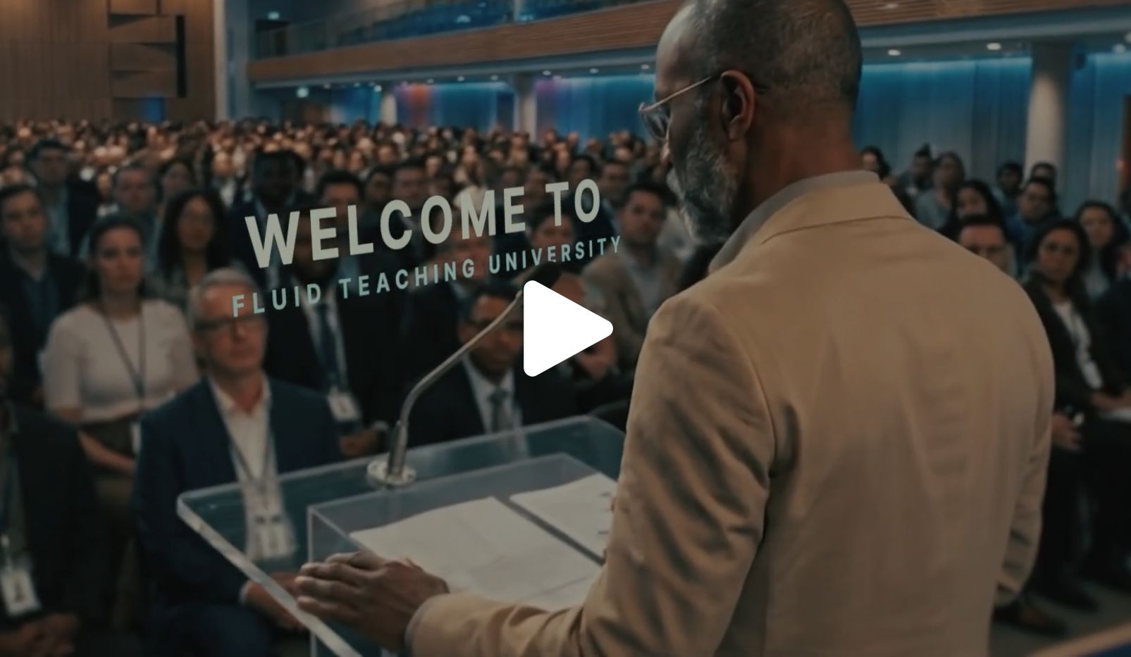 A man stands at a podium addressing a large audience in an auditorium. Words on screen read Welcome to Fluid Teaching University. A play button icon is overlaid at the center.